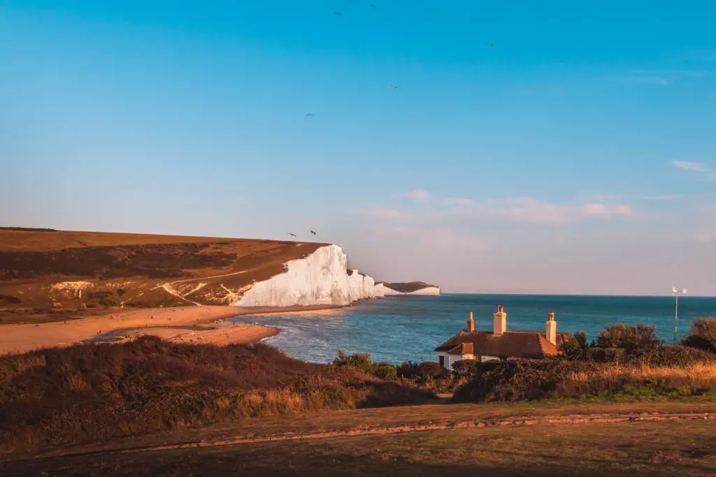 The white chalk seven sisters cliffs in the distance on the walk near Seaford.I The sea to the right of it is blue, and there is field with the coastguard cottage in the foreground.