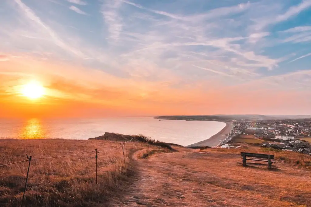 Standing on top of the hill, looking down at the curved coastline and buildings at the end of the walk from Southease to Seaford via Alfriston. The low hanging sun s on the left of the frame creating a warm, pink glow. There is a bench on the hill, looking down to the town.