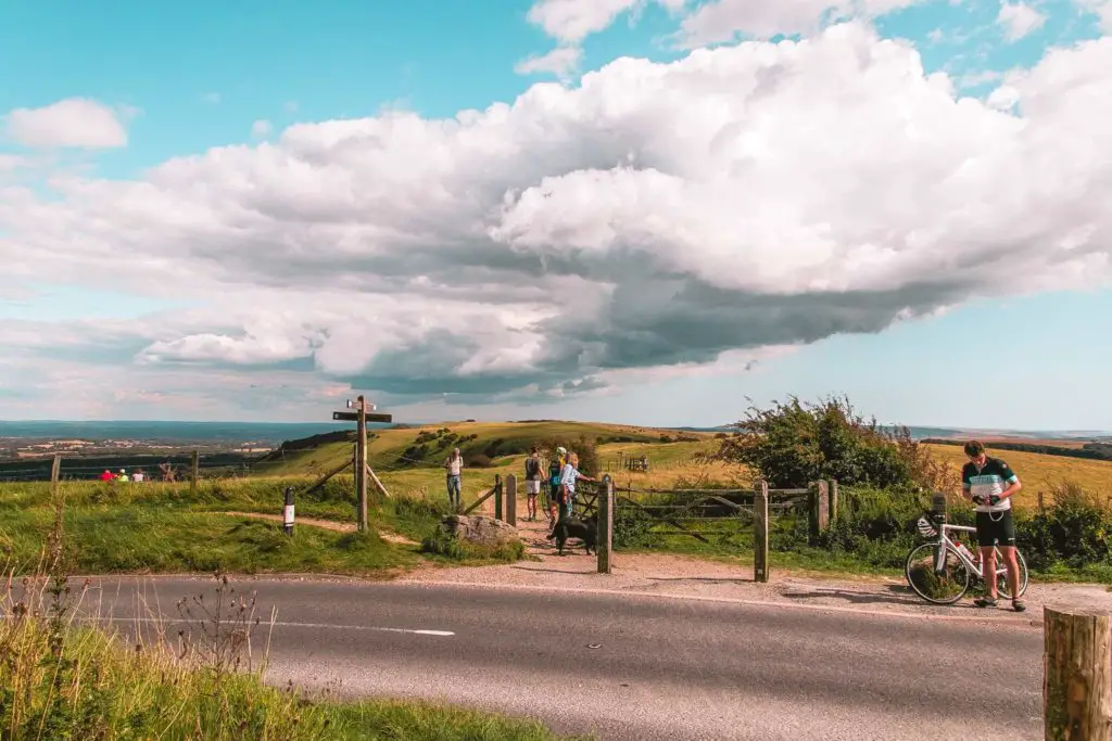 A road with a field on the other side on the walk to Ditchling Beacon. There is a man with his bike, and people walking in the field and through the gate.