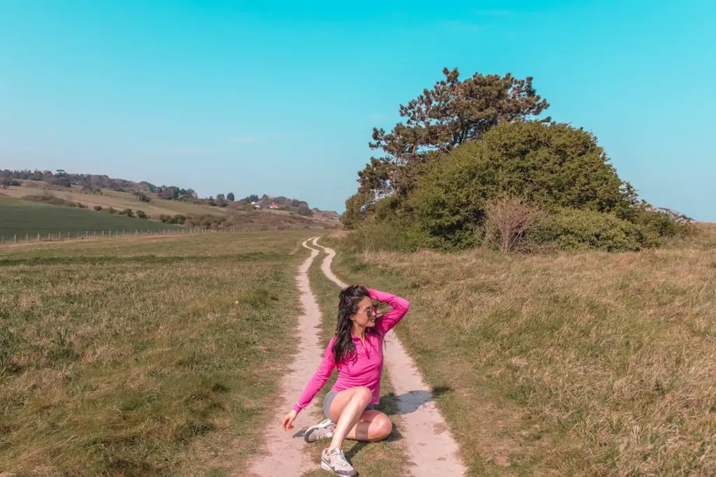 Zoe tehrani on the England Coast path from Dover to Deal. She is sitting on the path wearing a pink top, with green grass either side of the path and a blue sky with no clouds.