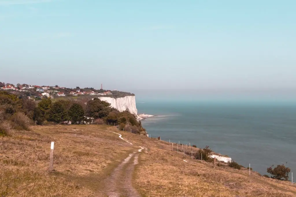 The England coast path trail with st Margaret's at Cliffe up ahead. You can see a bit of white cliffs rising from the English Channel. The sky is blue.