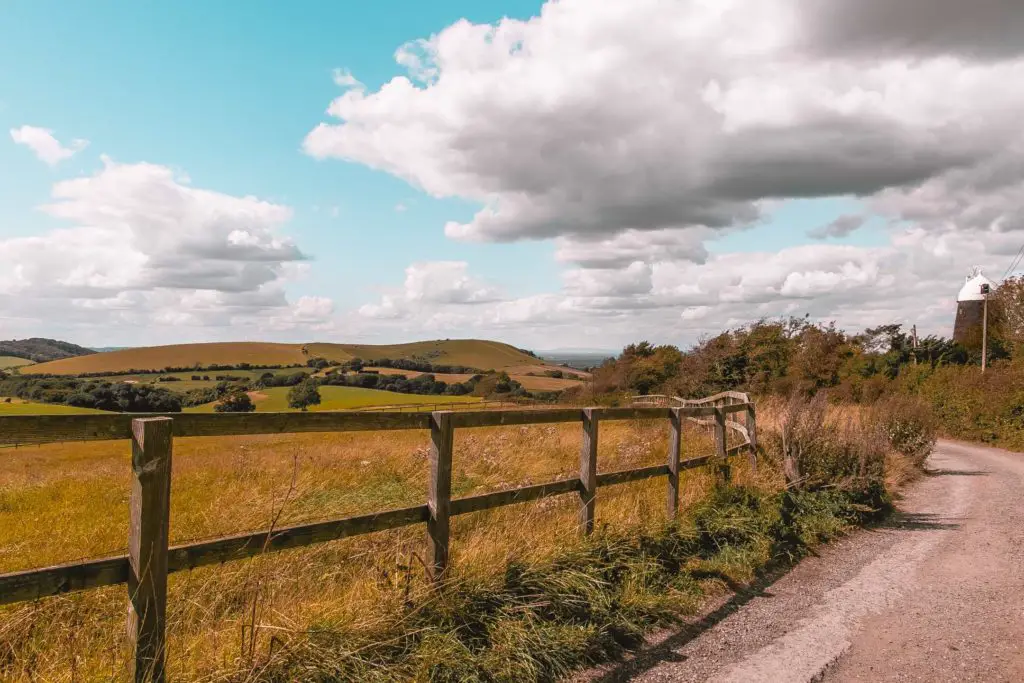 A path on the right separated to the field on the left by a fence on the walk from Hassocks to Lewes. The rolling South Downs hills are in the background. A black windmill oil visible in the right of the frame.