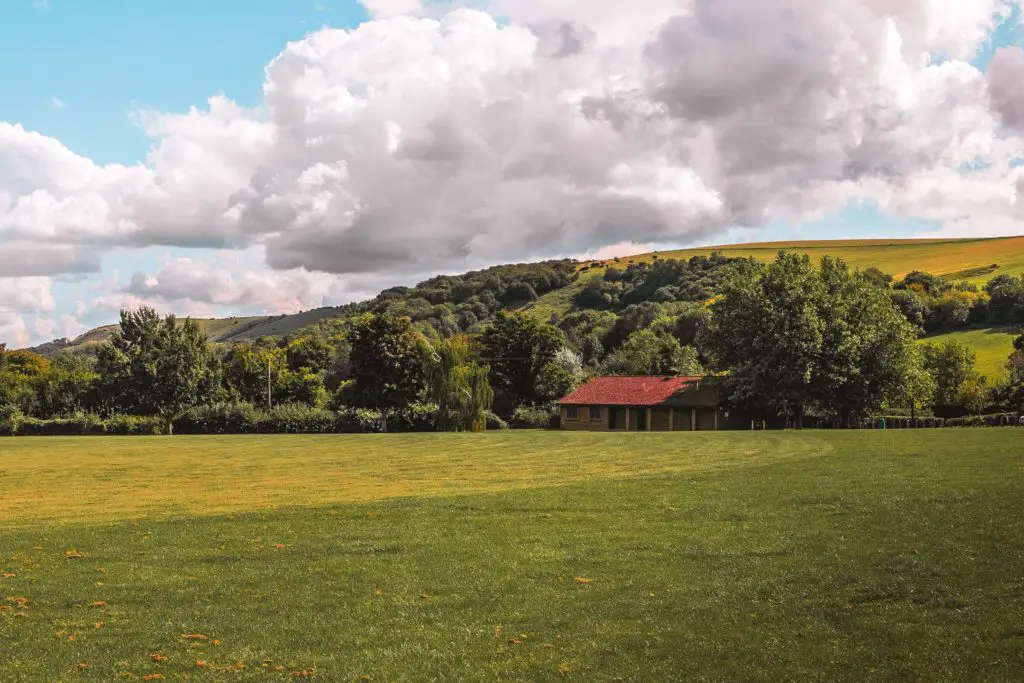 A green grass field with a red building on the other end on the walk out of Hassocks. There is a green and yellow hill in the background, and lots of trees.