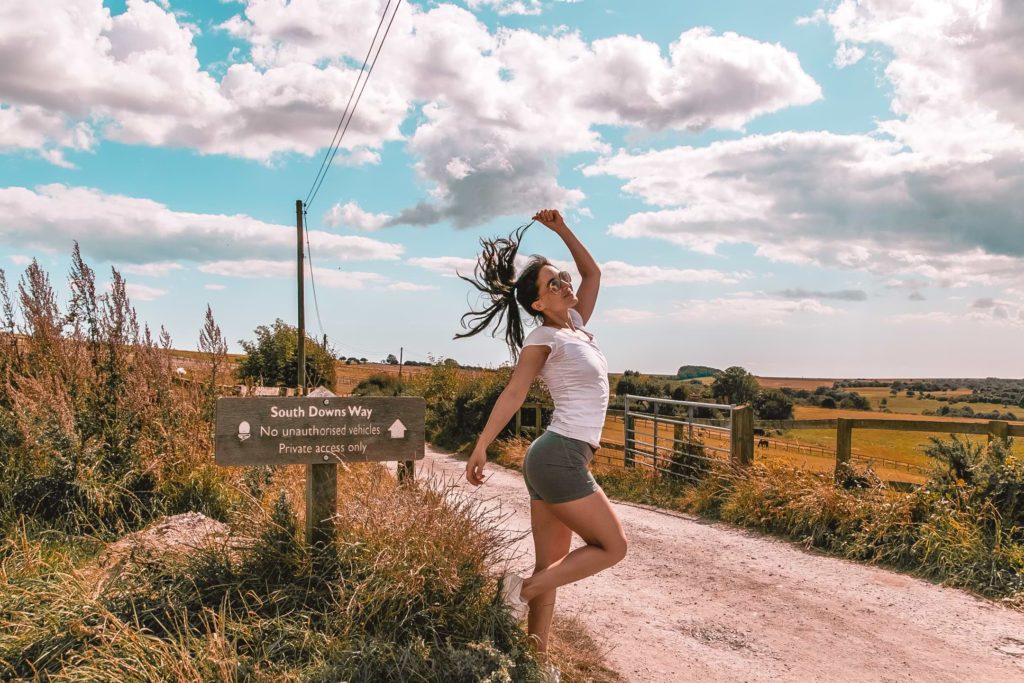 Zoe Tehrani posing next to South Downs Way signage. She is on a path with the South Downs fields and hills in the background.