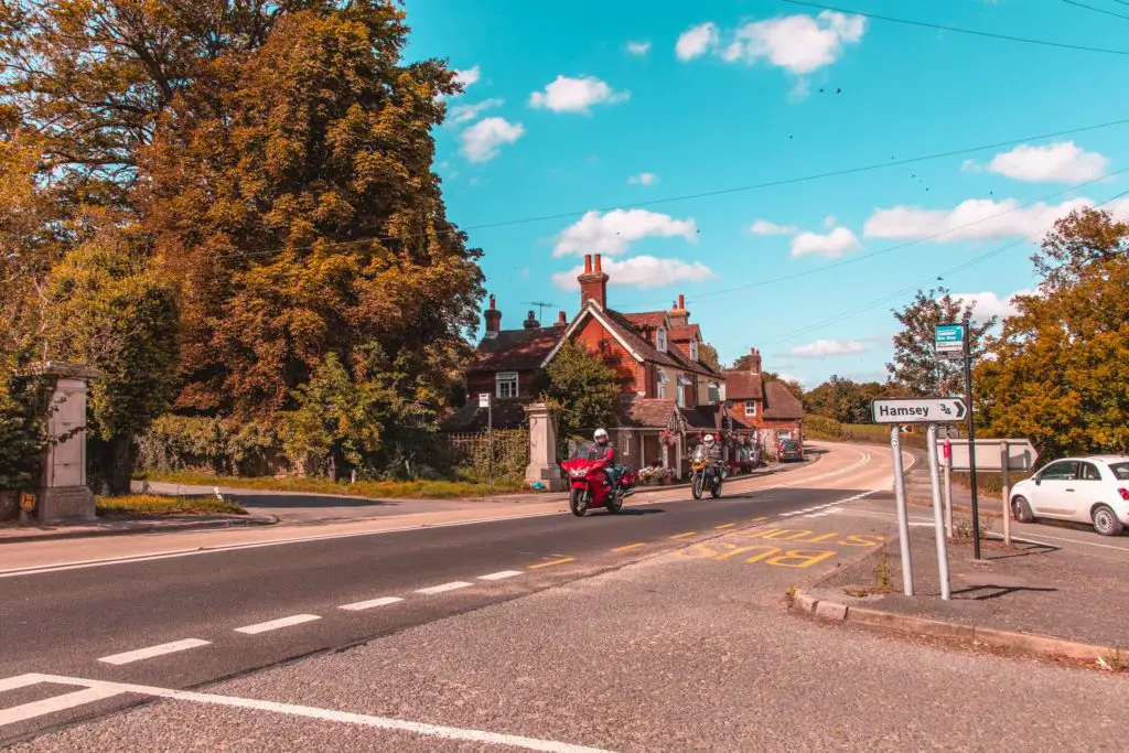 A road with two motorcyclists. There are houses on the other side of the road next to big trees and bushes. 