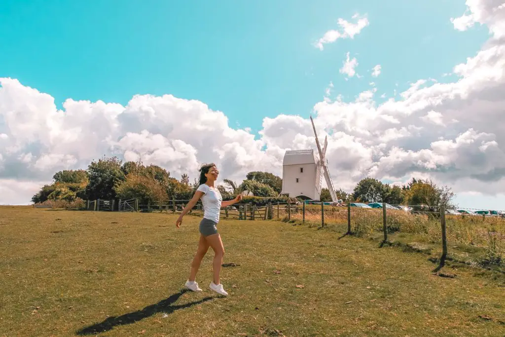 Zoe Tehrani standing on the green grass hill in front of a white windmill. The sky is blue with white fluffy clouds. 