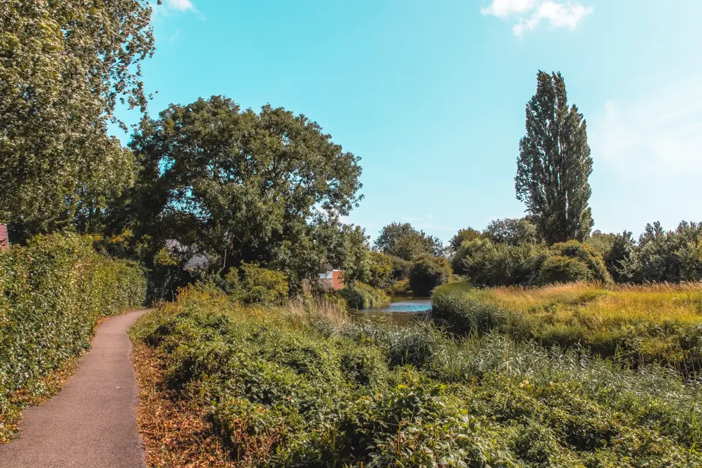 The path on the left with greenery to the right and a glimpse of the river. On a sunny day with blue sky.