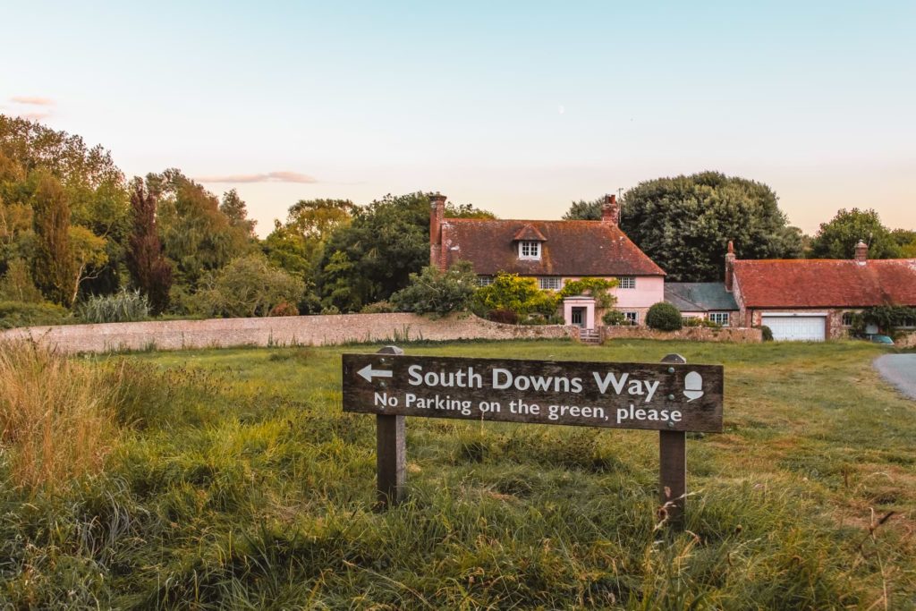 South Downs Way signage on green grass on the walk from Lewes to Southease. There are cute country cottages in the background, with trees behind them.