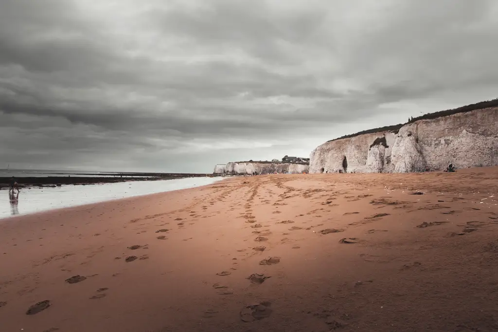 A long stretch of sand with footsteps. and white cliffs to the right, on one of the coastal walks near London. It is a grey moody day.