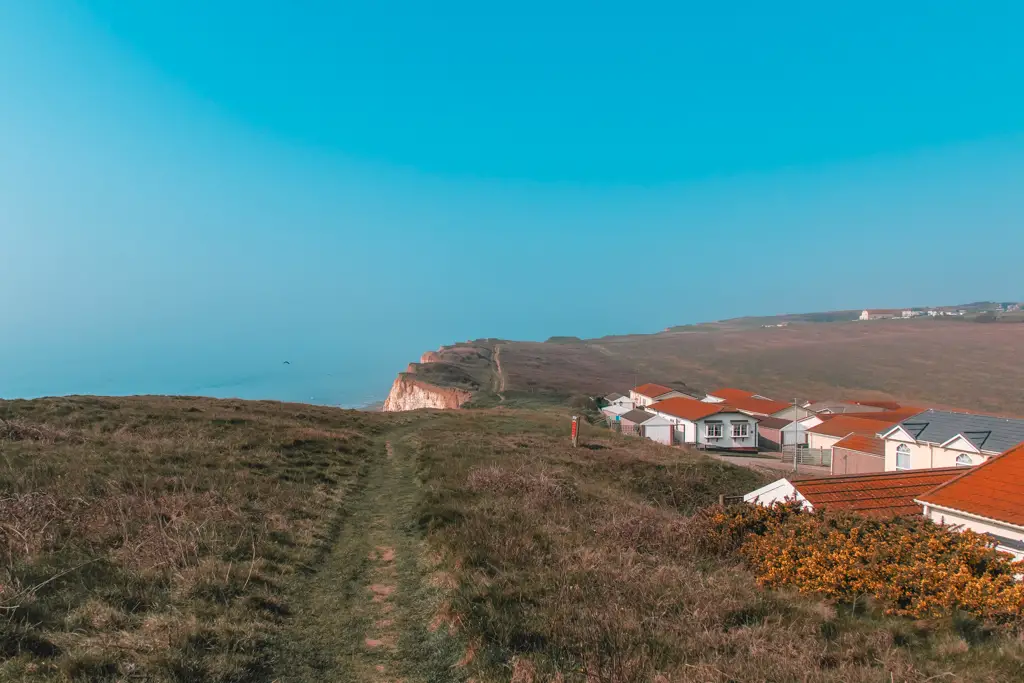 South East Coast path trail on the grassy clifftop. The sky is blue, and there are a few orange roofed homes on the right.