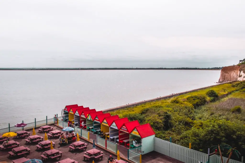 A view of a restaurant with colourful red roofed beach huts as seating areas. Next to the still ocean with a cliff view in the distance.