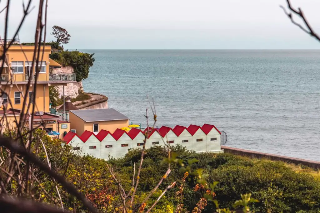 A view through an opening in the bushes looking at the red roofed huts next to the ocean.