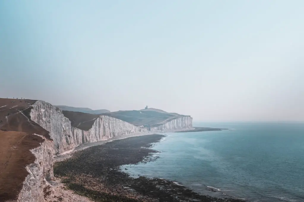 A view of the Seven Sisters white cliffs on the hike from Seaford to Eastbourne.It is a misty day. The sea is blue and Belle Tout lighthouse silhouette is visible in the distance on the cliff top.