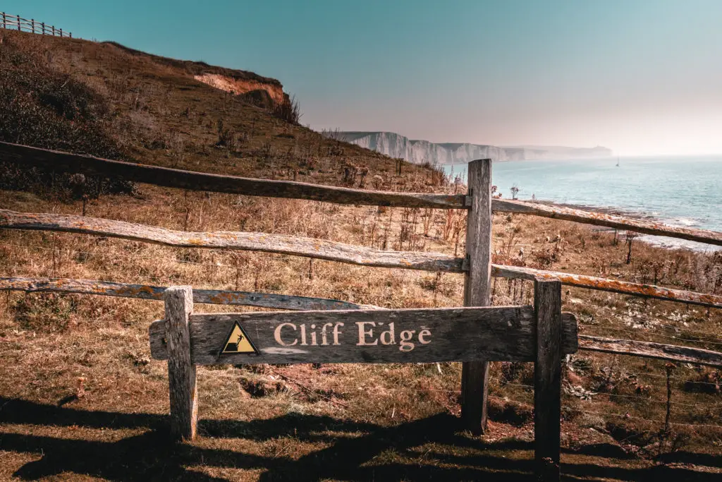 A wooden sign saying cliff edge with an image of someone falling off the cliff, on the seven sisters walk. The seven sisters cliffs are visible in the distance.