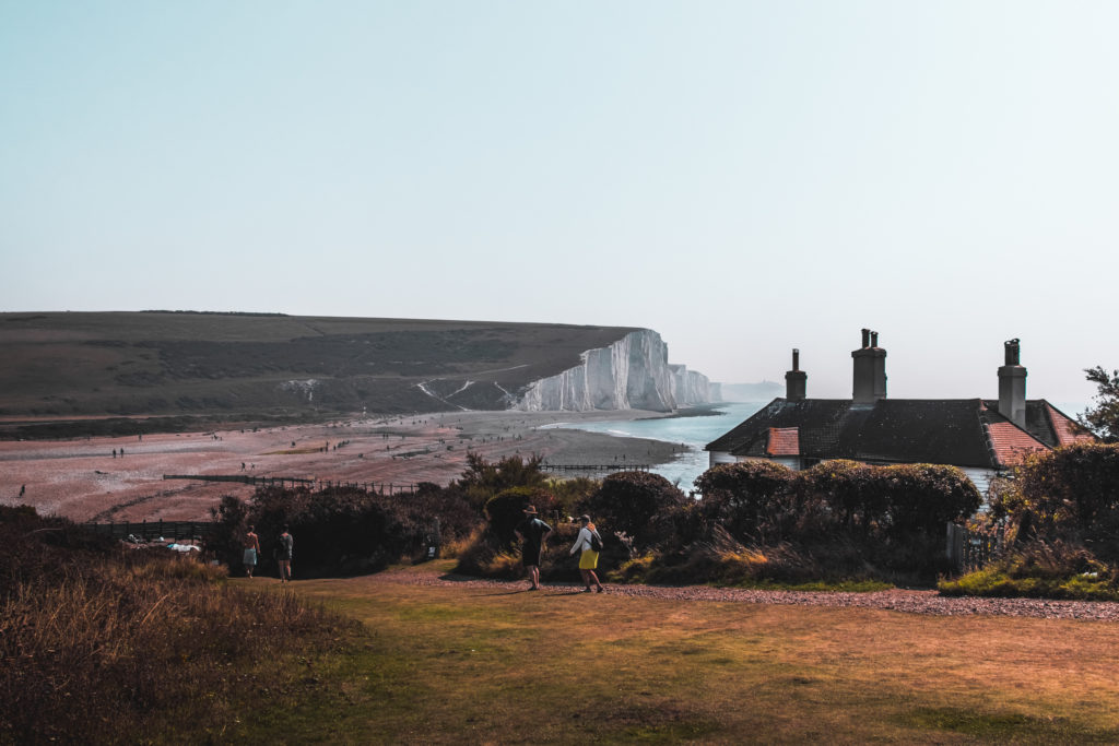 Entering Cuckmere Valley on the seven sisters cliffs walk. The cliffs are visible in the distance, there is a small house on the right of the grass path. The House is surrounded by bushes.