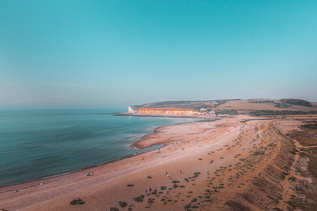 A hill top view of a stretch of beach and white cliffs in the distance. There are a few people on the beach. The sky and sea are blue.