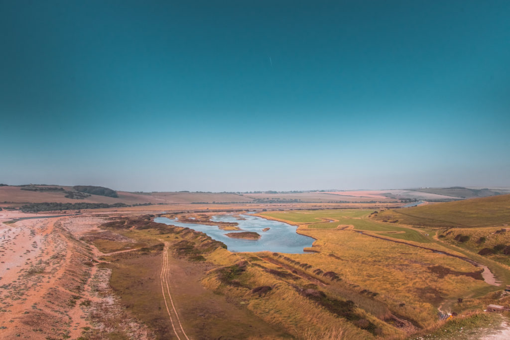 A view over the Cuckmere Valley. The ground is a mix of green and orange and the sky is blue.