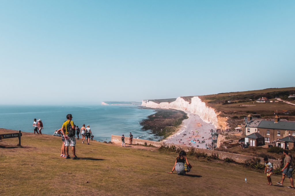 A view down over Birling Gap. The white cliffs next to a beach, with the visitors centre on the right. There are lots of people on the hill and on the beach.