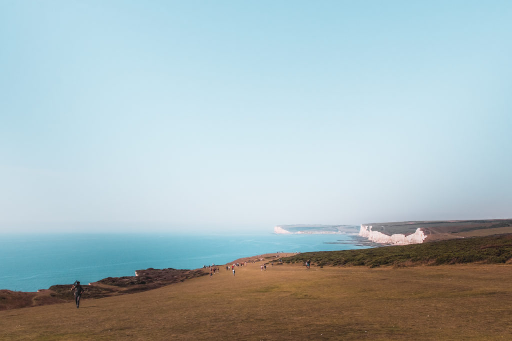 A view looking back on the Seven Sisters Cliffs hike. The clifftop landscape is green grass, with white cliffs views in the distance where they meet the blue sea.