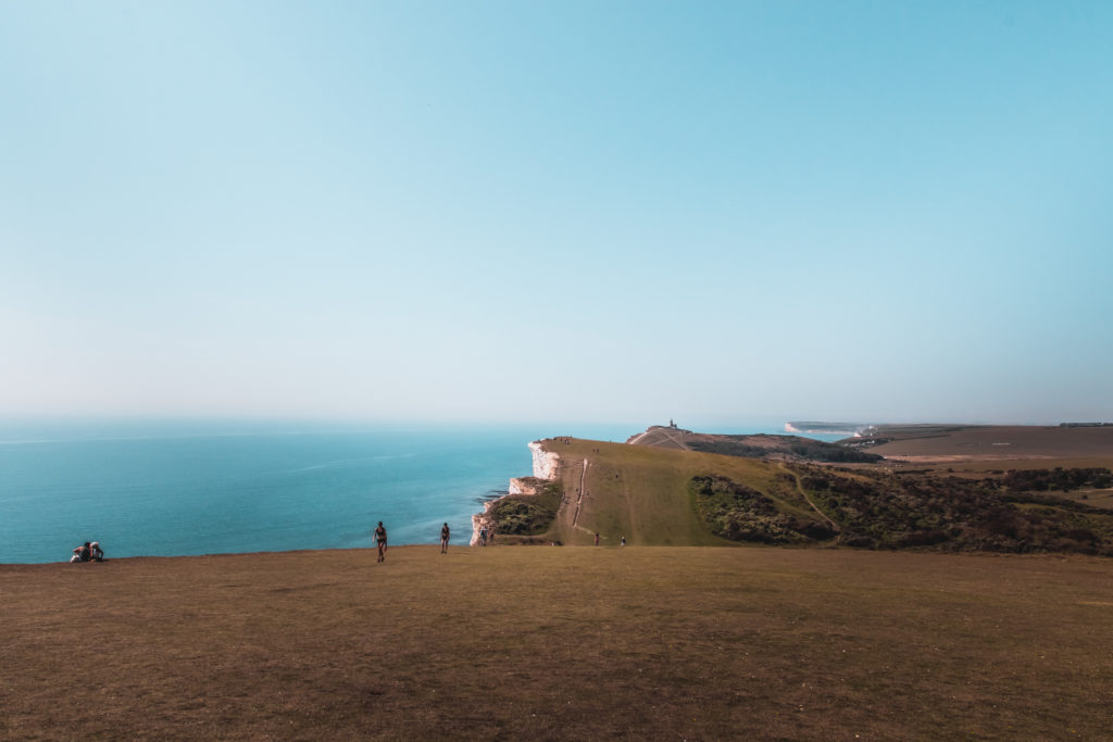 A view of the green grass clifftop landscape meeting the blue sea on the walk from Seaford to Eastbourne. There are a few people visible on the trail.