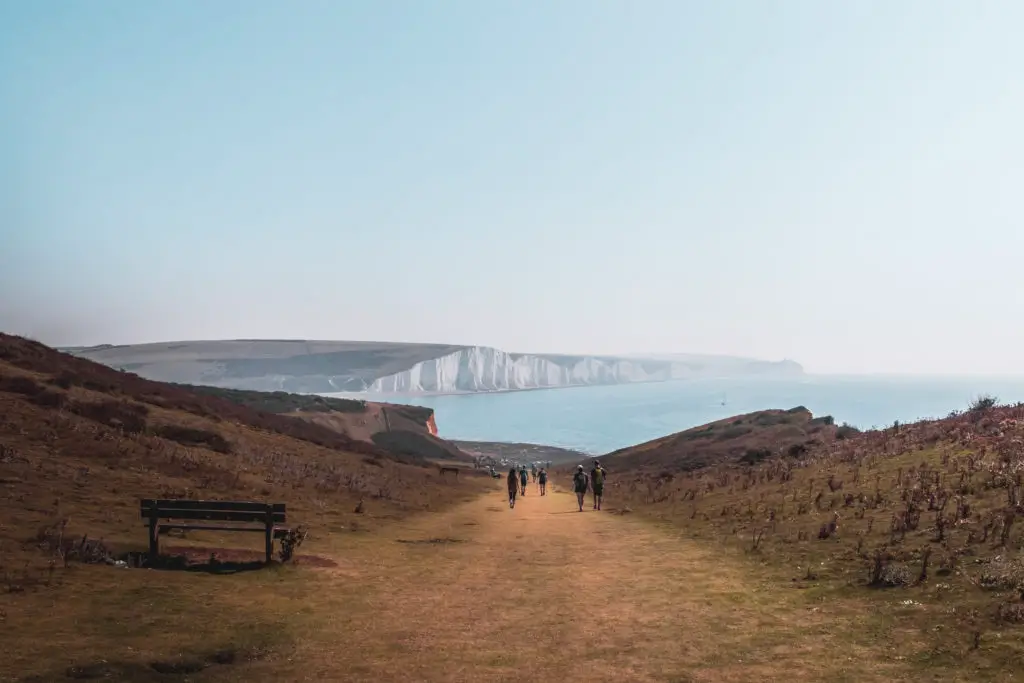 A view of the seven sisters cliffs on the walk from Seaford to Eastbourne. Walking along the grass trail. There is a bench on the left and some people ahead in the distance.