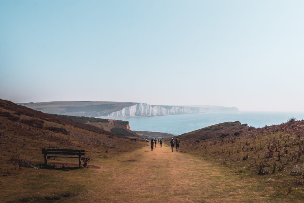 A view of the seven sisters cliffs on the walk from Seaford to Eastbourne. Walking along the grass trail. There is a bench on the left and some people ahead in the distance.