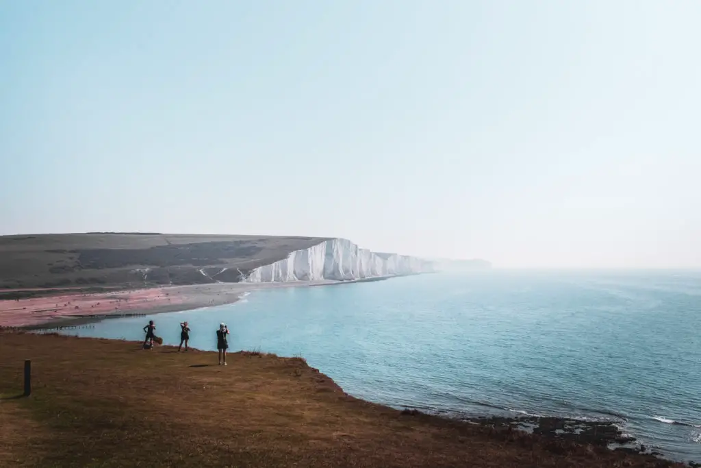 A view of the seven sisters cliffs on the walk from Seaford to Eastbourne. There are 3 people standing near the edge taking a photo. The sea and sky are blue.