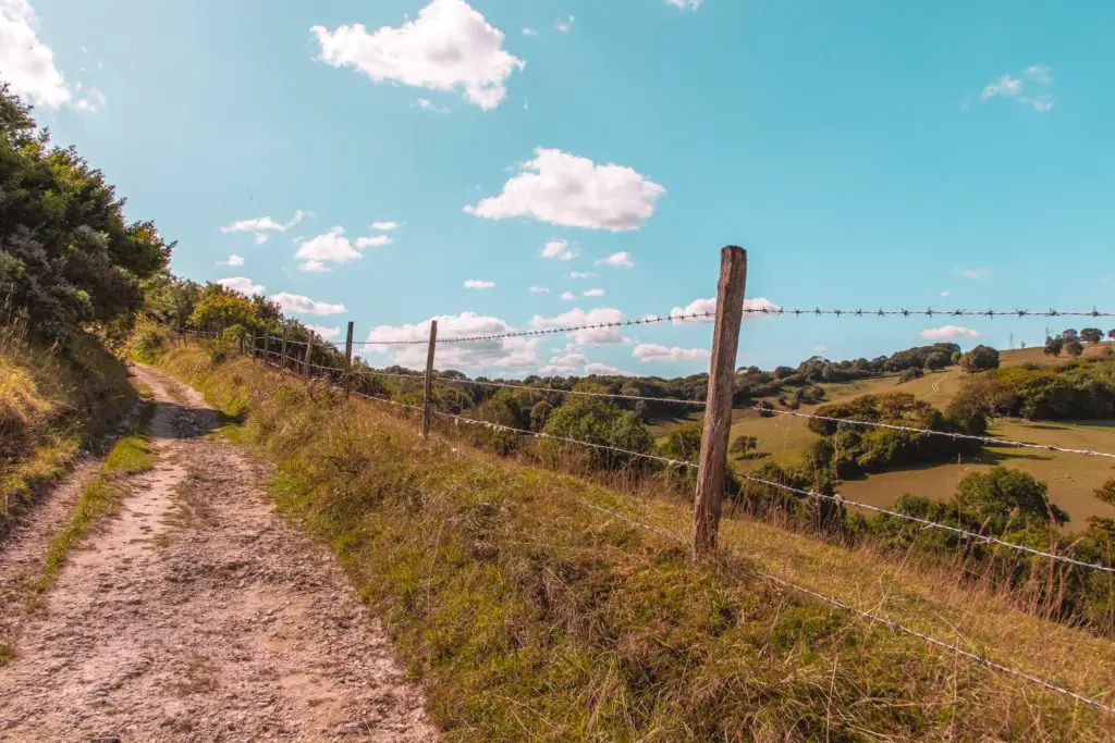 The uphill dirt trail on the left of the frame, with barbed wire fencing on the right when hiking near Lewes. There are green fields down below on the right with trees dotted about. The sky is blue with a few fluffy white clouds.