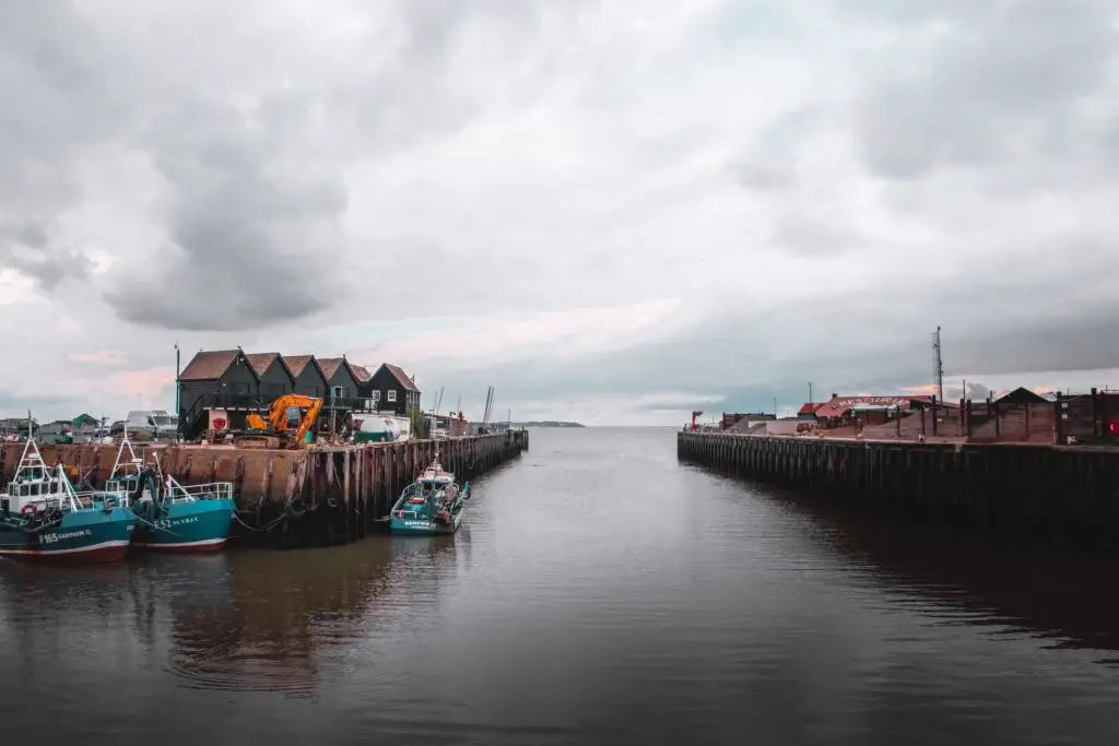 Whitstable Harbour, with a few blue coloured fishing boats on the black water. The sky is cloudy.