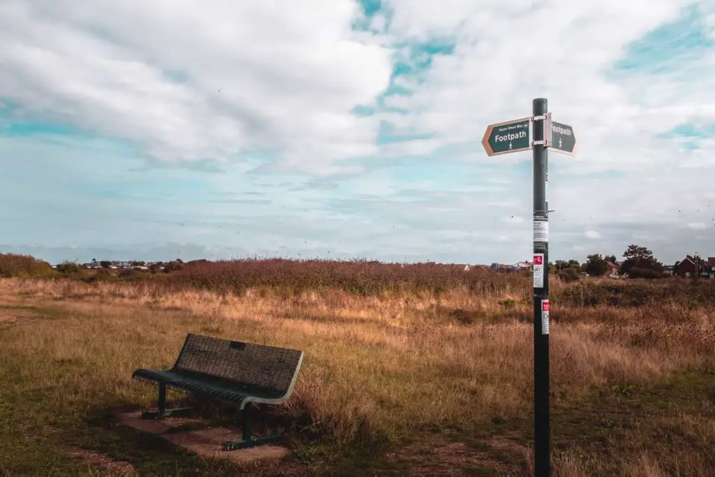 A bench next to footpath signage with the bird bush in the background. The sky is blue and mostly covered with white clouds.