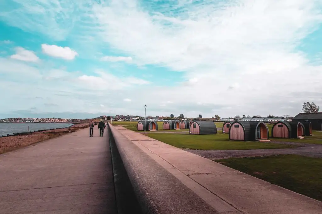 The coastal walking trail from whitstable to Herne Bay. There is a wall and on the other side is a green field with black camping pods. 