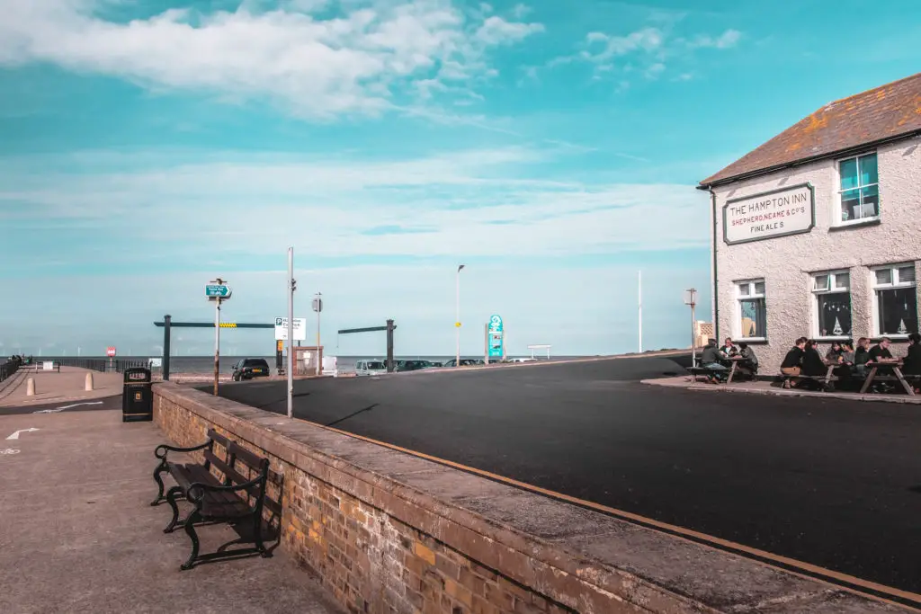 A path with a bench with its back to a wall against and elevated black tarmac road. There is a pub on the right, next to the roads with people sitting outside on benches. 