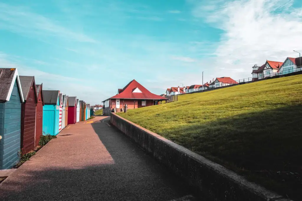 The coastal path in Herne Bay, with a green hill on the right side, and a row of beach huts on the left. The sky is blue.