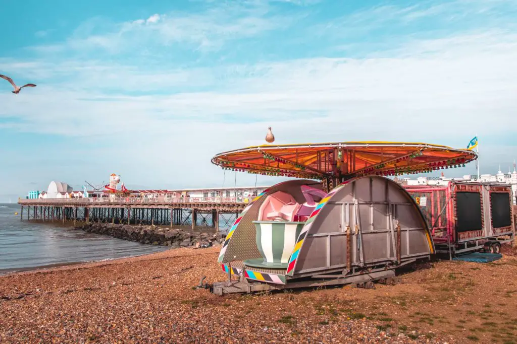 A tea cup ride closed up, on the shingle beach in Herne Bay. There is a seagull flying on the left, and the pier in the background over the sea.