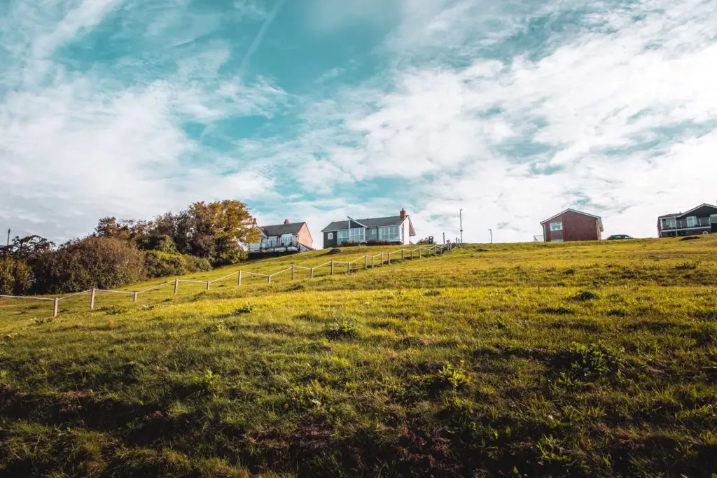 A green grass hill with a few houses on the top and a backdrop of blue sky with white streaky clouds.