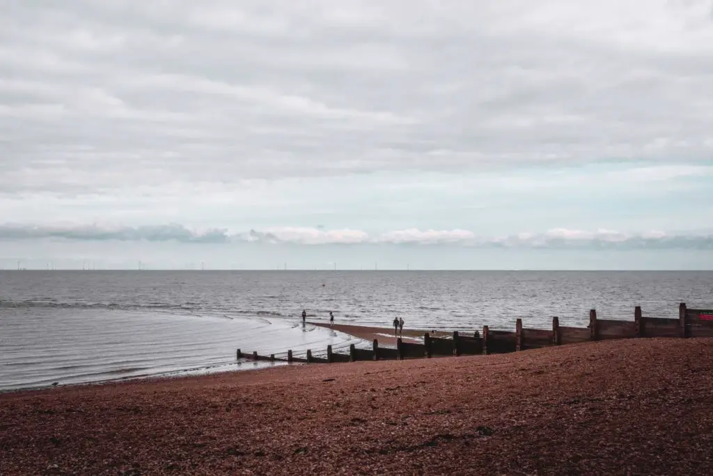 A shingle beach meeting the dark sea on the coastal walk from Whitstable to Herne Bay. There are a few people down by the sea.