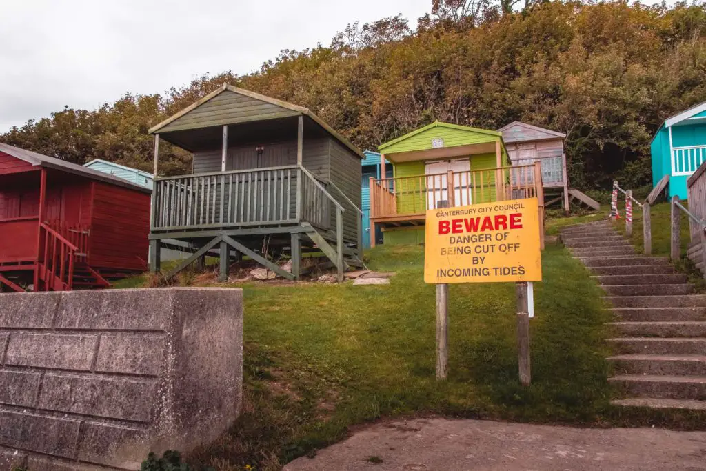 A group of beach huts in different colours on a hill with trees and bushes behind them. There is a yellow sign warning about the incoming tide.