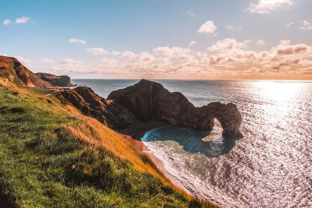 The rock arch formation of Durdle Door on the walk from Lulworth Cove. The grass is a mix of green and orange. The sky is blue with a hint of peach.