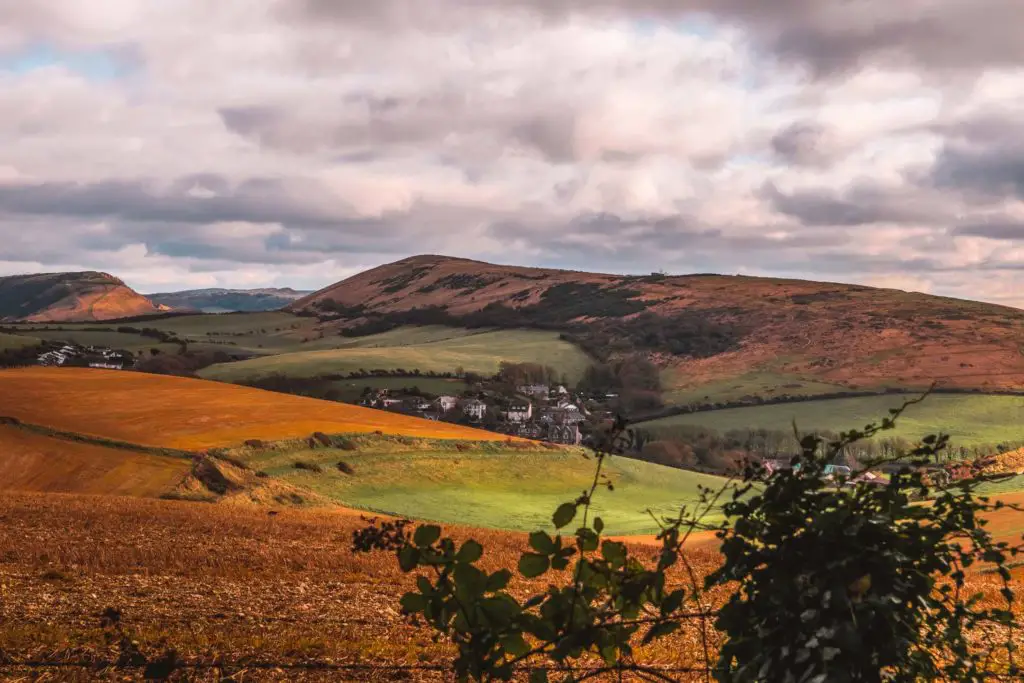 A view of a small cluster of houses surrounded by rolling hills in shades of green, o0range and red.