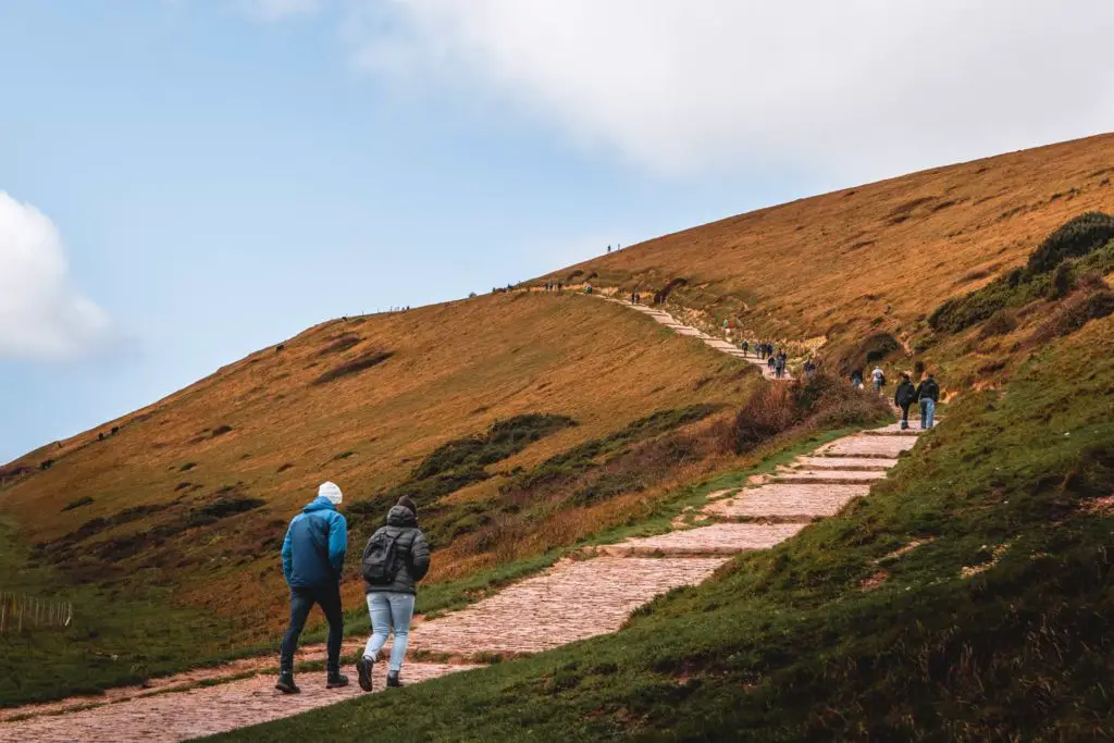 A trail leading up and over a hill on the walk from Lulworth Cove to Durdle Door. There are lots of people walking up it.