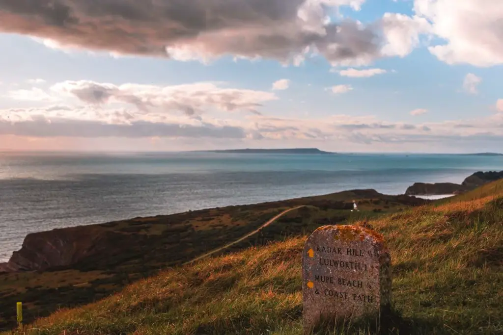 A slab of rock signage for Lulworth, Muoe Bay and the coast path. It is on top of a hill with a few of the sea and coastline below.