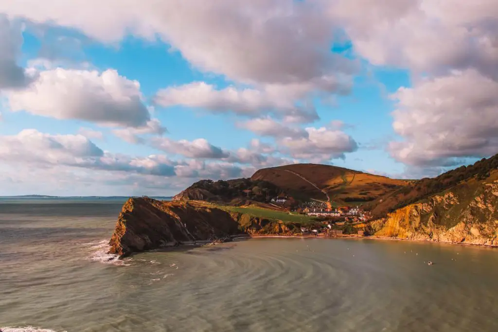 A view down to Lulworth cove. There are ripple in the sea. The sky is blue with white fluffy clouds.