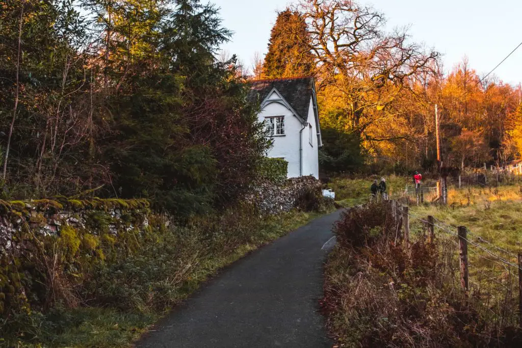 A road leading uphill past a white house on the left, surrounded by trees at the start of the walk from Ambleside to Elterwater and Loughrigg Tarn.