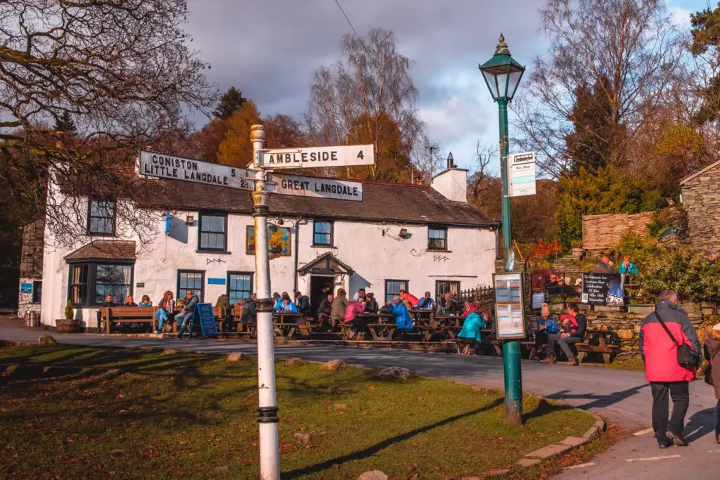 A white walled pub with lots of people sitting on the benches outside it. There is signage pointing to Ambleside, Great Langdale and Little Langdale.