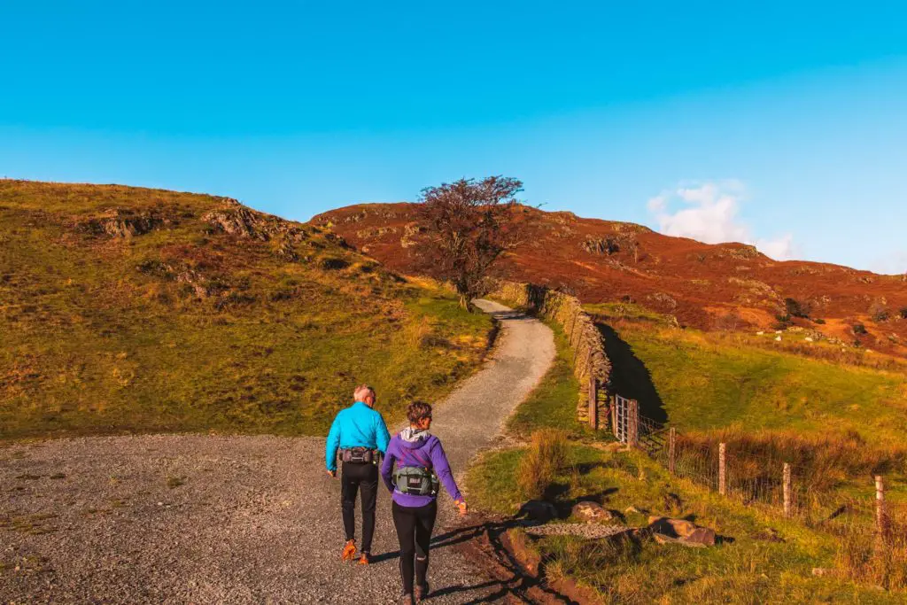 Two hikers on the trail leading away from Ambleside. The trail is surrounded by green and orange coloured fields and hills.