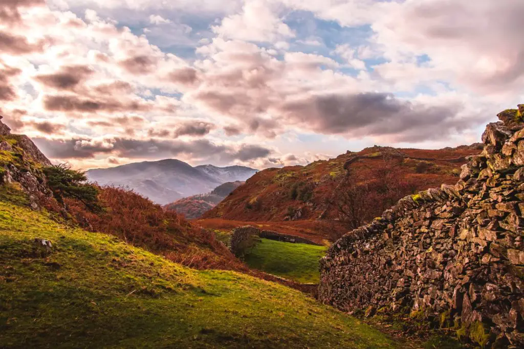 A grassy hill with a stone wall to the right and mountains in the distance on the circular walk from Ambleside to Elterwater.