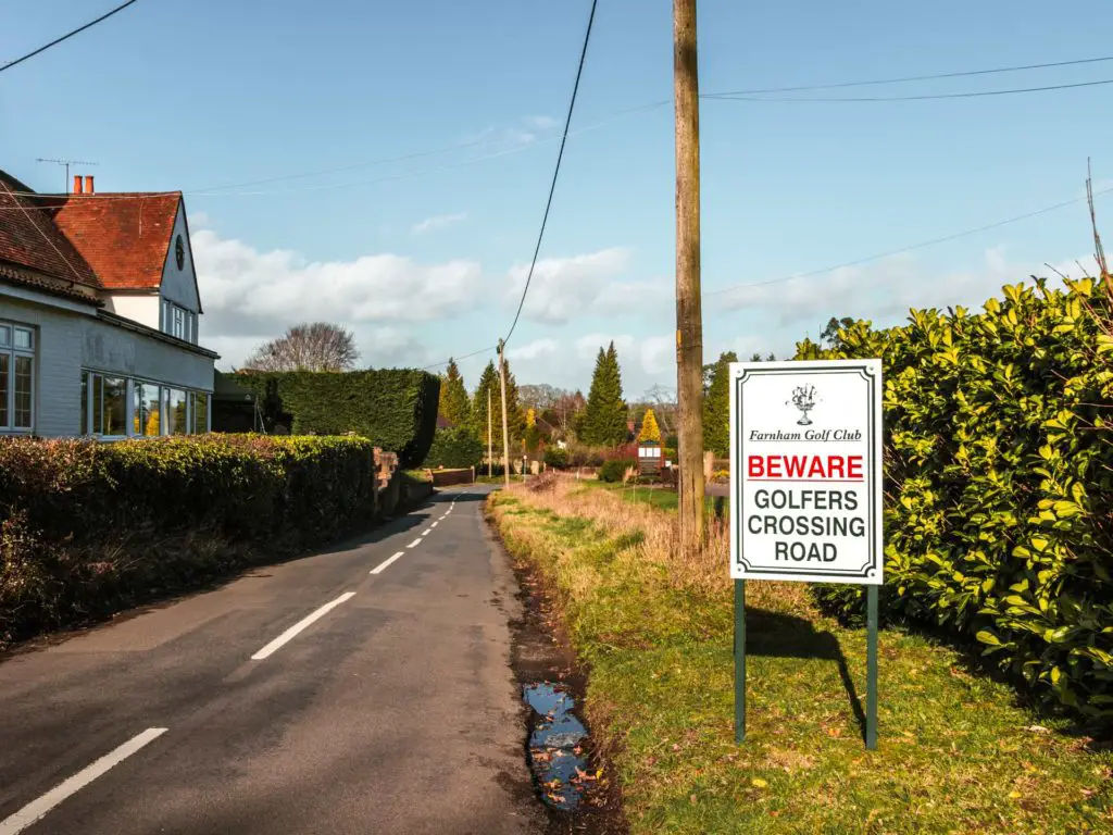 The main road, with a green and hedge on the right. There is a sign saying 'beware, golfers crossing road'. The sky is blue with a few clouds.