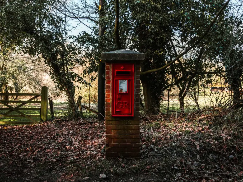 A brick post box under tree cover, on a ground covered on leaves on the walk from Farnham to Guildford.