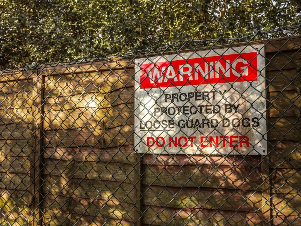 A wooden fence behind a metal fence with a sign warning of guard dogs.