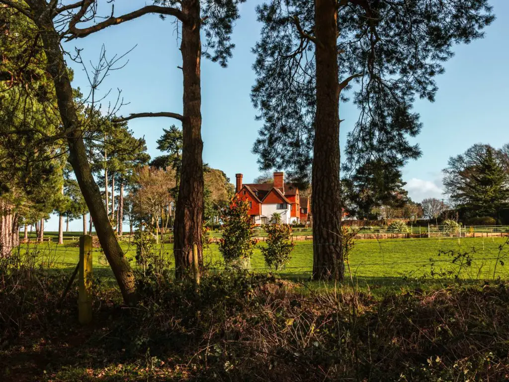 A house in the distance framed by the trunks of two trees whilst walking from Farnham to Guildford. Behind the trees and in front of the house is a well kept bright green Lawn.